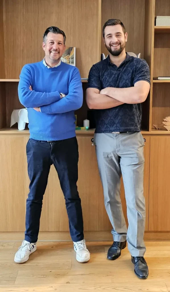 Two LaGrand team members standing side by side, smiling confidently, with their arms crossed in a modern office setting with wooden shelves in the background.